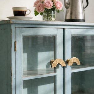 Może przedstawiać: Jasnoniebieska szafka ze szklanymi drzwiami i mosiężnymi uchwytami. Bukiet różowych róż, filiżanka kawy i srebrny czajnik stoją na górze. Szafka ma postarzaną powierzchnię.