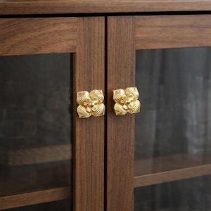 May include: A close-up of a wooden cabinet with two doors. Each door has a gold-colored, flower-shaped handle. The cabinet is a dark brown color. A woven basket sits on top of the cabinet.