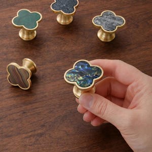 Puede incluir: Pomos decorativos para armarios con diseño de trébol de cuatro hojas. Los pomos tienen una base de latón y están rematados con diversos materiales, como piedra verde, piedra oscura y concha de abulón. Una mano sostiene un pomo.