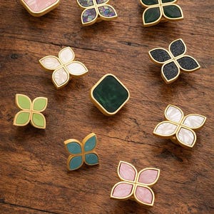 May include: A selection of decorative cabinet knobs in floral and geometric shapes. The knobs have gold-coloured metal frames with inserts in pink, green, blue, white, and black. They are displayed on a wooden surface.
