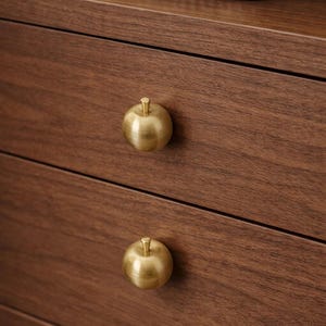 Op de afbeelding: Close-up van een houten dressoir met twee lades. Elke lade heeft een goudkleurige knop in de vorm van een appel. Het hout heeft een rijke, donkerbruine kleur en zichtbare nerf. Bovenop het dressoir staat een gevlochten mand.