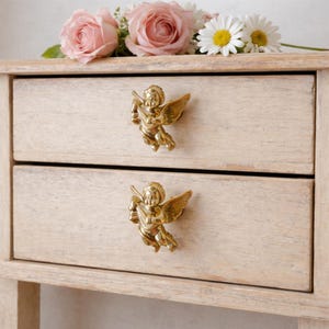 May include: A light-colored wooden nightstand with two drawers, each adorned with a gold-colored cherub-shaped handle. Pink roses and white daisies rest on top of the nightstand, adding a touch of floral elegance.