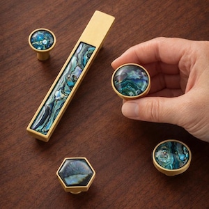 May include: A collection of decorative cabinet hardware featuring brass finishes and abalone shell inlays. The set includes a long pull, round knobs, and a hexagonal knob. The abalone shell displays iridescent blues, greens, and purples.