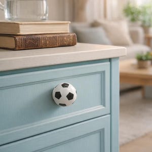 Może przedstawiać: Jasnoniebieska szafka z gałką w kształcie piłki nożnej. Szklany czajnik i dwie książki znajdują się na szafce. Gałka piłki nożnej jest biała z czarnymi pięciokątami. Szafka jest w pokoju z lampą i sofą.