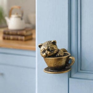 May include: A bronze cat door knob in the shape of a sleeping cat in a teacup. The door knob is mounted on a light blue door. A white teapot and books are visible in the background. The teacup is a decorative element.