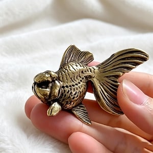 Op de afbeelding: Een gedetailleerd goudvisbeeldje in messingkleur. De sculptuur heeft getextureerde vinnen en een afgerond lichaam, met ingewikkelde details. De vis wordt in de hand van een persoon gehouden, wat de grootte en het vakmanschap benadrukt. Dit decoratieve item is geschikt voor woondecoratie.