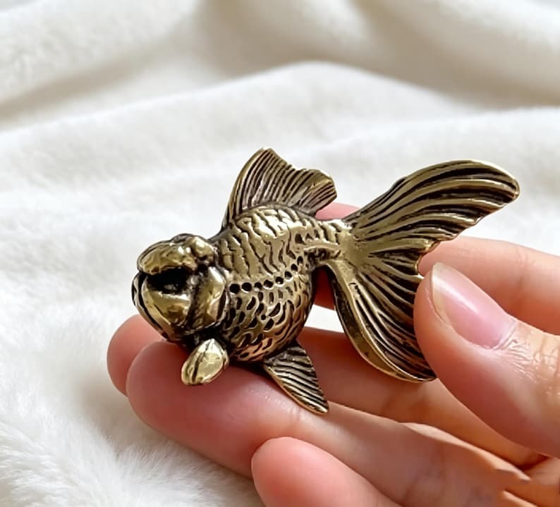 Op de afbeelding: Een gedetailleerd goudvisbeeldje in messingkleur. De sculptuur heeft getextureerde vinnen en een afgerond lichaam, met ingewikkelde details. De vis wordt in de hand van een persoon gehouden, wat de grootte en het vakmanschap benadrukt. Dit decoratieve item is geschikt voor woondecoratie.