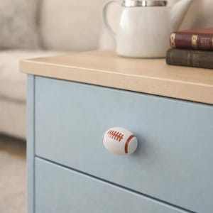 Könnte beinhalten: Ein hellblauer Schrank mit einem Baseball-Knauf. Der Knauf ist weiß mit roten Nähten und Akzenten. Eine weiße Teekanne und Bücher befinden sich auf der hellen Holzoberfläche.
