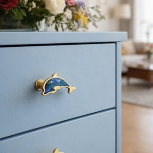 May include: A light blue dresser with two drawers, each adorned with a dolphin-shaped knob. The knobs are gold-colored with a blue and green iridescent inlay. A vase of colorful flowers sits on top of the dresser.