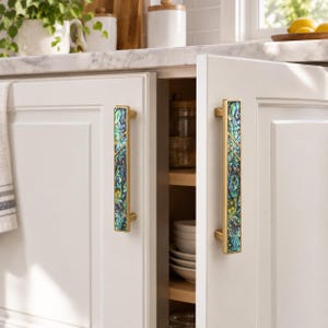May include: Two vertical cabinet pulls with a brushed gold metal frame and a central inlay of iridescent abalone shell featuring swirling patterns of blue, green, and purple. The handles are mounted on white cabinet doors with a recessed panel.