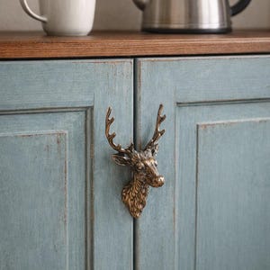 Könnte beinhalten: Nahaufnahme eines blauen Schranks mit einem Hirschkopfgriff aus Messing. Eine weiße Tasse und ein silberner Wasserkocher stehen auf einer Holzplatte über dem Schrank. Die Schranktüren haben ein Used-Finish.