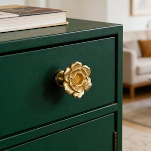 May include: A close-up of a dark green cabinet with a gold-colored flower-shaped drawer pull. The drawer pull is centered on the drawer and has detailed petals. A book is on top of the cabinet.