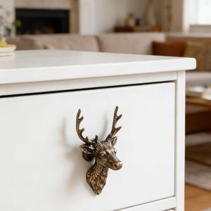 May include: A bronze-coloured deer head drawer pull mounted on a white cabinet. The deer head is detailed with antlers and facial features. The cabinet has two drawers and is in a room with a fireplace and sofa.