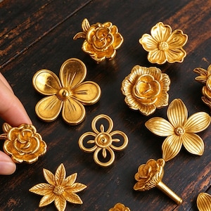 May include: A collection of gold-colored metal floral and geometric decorative knobs. The set includes various flower shapes, including roses and five-petaled blossoms, alongside a symmetrical four-loop design, all featuring a metallic finish.