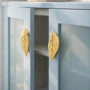 May include: A light blue cabinet with glass doors and gold leaf-shaped handles. The cabinet doors are slightly ajar, revealing a white interior. Two books are on top of the cabinet. The cabinet is a piece of furniture.