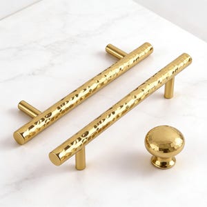 May include: Two gold-colored, hammered-texture cabinet pulls and a matching knob. The pulls are bar-shaped with cylindrical ends. The knob is spherical with a short, cylindrical base. All pieces have a bright, reflective finish.