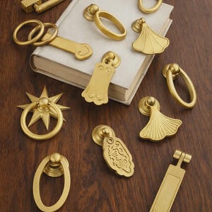 May include: A collection of brass cabinet hardware, including ring pulls, starburst pulls, and rectangular pulls. The hardware is displayed on a wooden surface and a book.