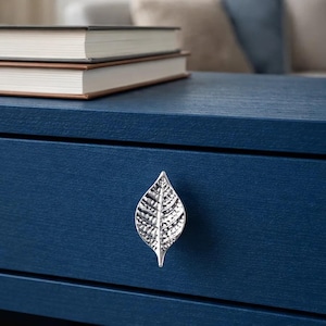 Pode incluir: Uma gaveta azul marinho com uma puxador prateado em forma de folha. Dois livros com capas castanhas e pretas estão empilhados em cima. A gaveta tem um acabamento texturizado e o puxador é detalhado.