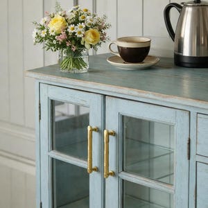 May include: A light blue cabinet with glass doors and brass handles. A bouquet of yellow roses and daisies sits in a glass vase. A cup of coffee and a stainless steel kettle are on top.