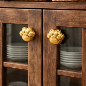 Könnte beinhalten: Ein dunkelbrauner Holzschrank mit Glastüren und goldenen pfotenförmigen Knöpfen. Im Schrank befinden sich weiße Teller. Oben sind Weidenkörbe zu sehen. Rustikales Design.