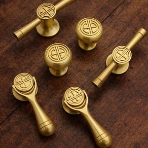 May include: A collection of brass cabinet hardware, including knobs and pulls. The hardware features a circular design with an embossed pattern. The pieces are arranged on a dark wood surface, showcasing their warm, metallic finish.
