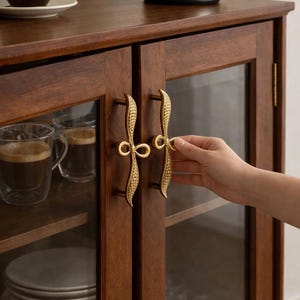 Puede incluir: Un mueble de madera oscura con puertas de cristal y tiradores dorados decorativos. En el interior, tazas de cristal con café y una pila de platos. Una tetera plateada está encima. Los tiradores son una característica clave.