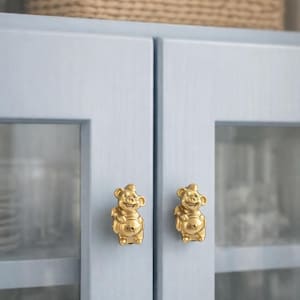 May include: Two gold-colored cabinet knobs shaped like smiling bears. The knobs are mounted on a light blue cabinet door with glass panels. A woven basket sits on top of the cabinet.