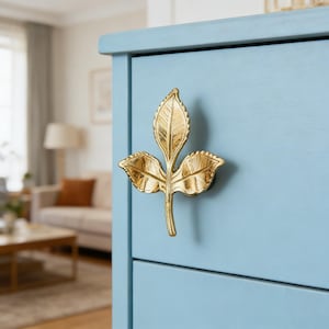 May include: A gold-colored leaf-shaped cabinet pull is mounted on a light blue cabinet. The pull features three detailed leaves and a stem. The cabinet has a simple design and is painted in a soft, pastel blue color. The pull adds a touch of elegance.