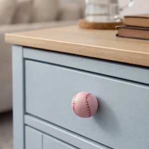 Op de afbeelding: Een lichtblauwe kast met een knop in de vorm van een honkbal. De knop is roze met rode stiksels, die op een honkbal lijken. De kast heeft een licht houten blad. Een glazen kan en boeken zijn zichtbaar.