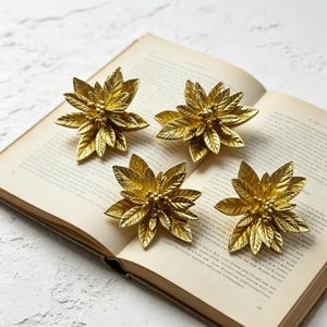 Puede incluir: Cuatro objetos decorativos dorados con forma de flor, con diseños detallados de hojas y cuentas. Están dispuestos sobre las páginas abiertas de un libro antiguo, creando una estética cálida. Los objetos parecen ser de metal.
