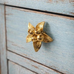 May include: A gold-colored floral cabinet knob with a cluster of berries. The knob is mounted on a light blue, distressed wooden cabinet door. The knob's design features leaves and berries.