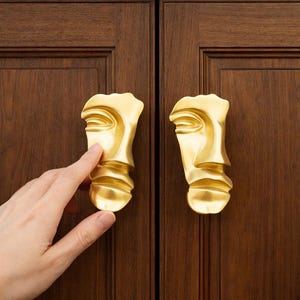 Puede incluir: Dos tiradores dorados para armario con forma de rostro estilizado. Los tiradores están montados en una puerta de armario de madera marrón oscuro. Una mano está tocando uno de los tiradores.