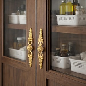 以下が含まれることがあります： ガラス扉と装飾的な金色の取っ手が付いた木製キャビネット。キャビネット内には、白いプラスチック製のバスケットに整理されたさまざまなボトルや瓶が並んでいます。木材は濃い茶色です。