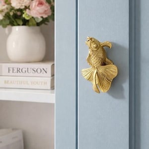 May include: A gold-colored fish-shaped cabinet knob mounted on a light blue cabinet door. The knob is detailed with scales and fins. A white vase with pink flowers is visible on a shelf in the background.