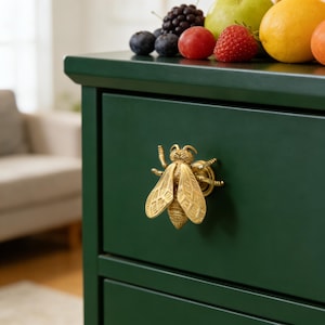 Puede incluir: Primer plano de un cajón de cómoda verde oscuro con un tirador dorado en forma de abeja. La parte superior de la cómoda está adornada con fruta fresca, como arándanos, fresas y un limón. El cajón está cerrado.