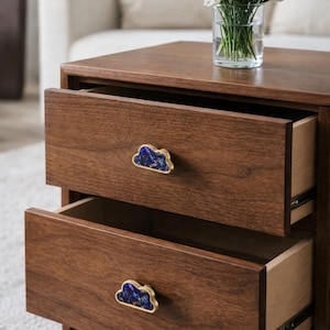 May include: A two-drawer wooden bedside table with cloud-shaped handles in blue and gold. The drawers are open, showing the interior. A vase of white flowers sits on top of the table. The table is made of wood.