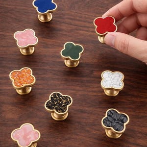 Pode incluir: Uma coleção de puxadores decorativos para armários em várias cores, incluindo azul, vermelho, rosa, verde, laranja, preto, branco e preto. Cada puxador apresenta um design de trevo de quatro folhas com uma base dourada. Os puxadores são exibidos em uma superfície de madeira.