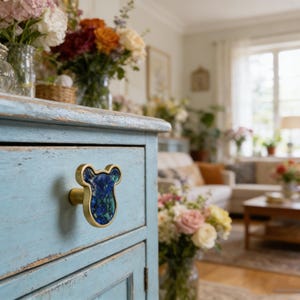 May include: A light blue painted wooden cabinet with a decorative brass drawer pull. The drawer pull has a blue and green stone inlay. The cabinet is in a room with flowers in vases.