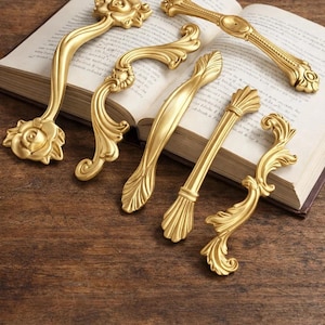 Puede incluir: Seis tiradores de gabinete dorados ornamentados con varios diseños decorativos, incluyendo patrones florales y de volutas. El hardware está dispuesto sobre un libro abierto con páginas envejecidas, sobre una superficie de madera oscura.