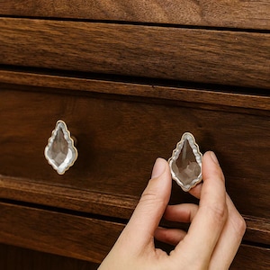 May include: Clear crystal cabinet knobs with a gold-coloured trim. The knobs are teardrop-shaped and mounted on a dark brown wooden cabinet. One hand is shown holding a knob.