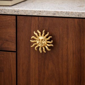 Op de afbeelding: Een goudkleurige, zonvormige kastknop met een lachend gezicht is gemonteerd op een donkerbruine houten kastdeur. De zon heeft gedetailleerde stralen en gelaatstrekken. De knop voegt een decoratief tintje toe aan de kasten.