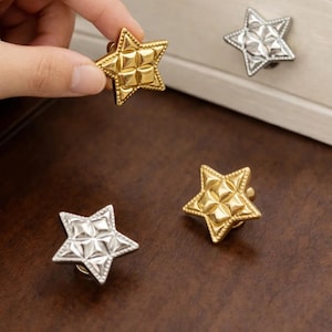 Può includere: Pomelli per mobili a forma di stella, dorati e argentati. I pomelli dorati hanno una superficie strutturata e un bordo decorativo. I pomelli argentati hanno un motivo geometrico. Una mano tiene un pomello dorato. Questi pomelli sono per mobili.