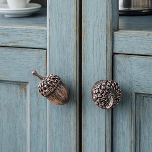 May include: Close-up of two acorn-shaped cabinet knobs. The knobs are bronze-colored with detailed textures, mounted on a light blue cabinet door. The cabinet has a distressed, rustic finish, suggesting a vintage or farmhouse style.