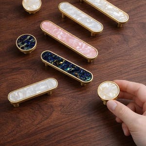 Pode incluir: Ferragens decorativas para armários em vários designs. Inclui puxadores redondos e puxadores retangulares com bases douradas. As inserções apresentam padrões iridescentes brancos, rosa e azul escuro. Uma mão segura um dos puxadores.