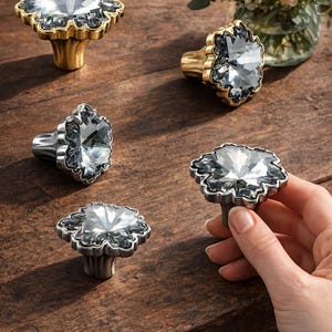Puede incluir: Pomos decorativos para armarios con centros facetados similares a cristales y bases metálicas ornamentadas. Algunos pomos tienen bases doradas, otros plateadas. Los pomos están dispuestos sobre una superficie de madera oscura, con un ramo de flores al fondo.