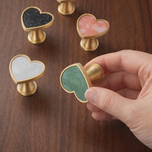 Puede incluir: Pomos de armario en forma de corazón en varios colores, incluyendo blanco, negro y rosa, con bases doradas. Un pomo se sostiene en una mano. Los pomos están hechos de un material sólido y están diseñados para muebles.