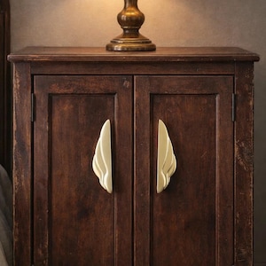 Puede incluir: Un mueble de madera marrón oscuro con dos puertas y tiradores dorados en forma de ala. Una lámpara de latón con una pantalla beige se encuentra encima del mueble. El mueble forma parte de un juego de dormitorio.