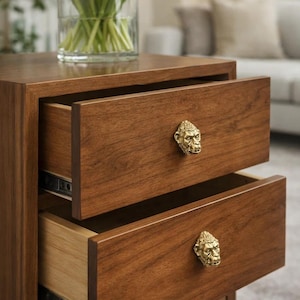 May include: A two-drawer wooden bedside table with brass gorilla face handles. The drawers are open, showing the interior. A vase of white tulips sits on top. The table is a warm brown colour.
