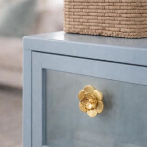 May include: A pale blue cabinet with a gold flower-shaped handle. A woven basket rests on top. The cabinet's design is simple and elegant, suitable for a bedroom or living room. The handle is a decorative feature.