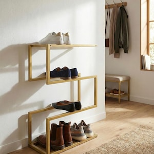 Op de afbeelding: Een moderne schoenenrek van goudkleurig metaal met een uniek zigzagontwerp. Het rek biedt plaats aan verschillende paren schoenen, waaronder sneakers, loafers en laarzen. Het is aan een witte muur bevestigd en creëert een stijlvolle opbergoplossing.